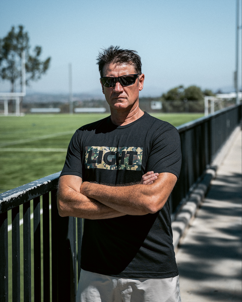 Man wearing sunglasses and a black t-shirt with a logo, standing on a sports field.