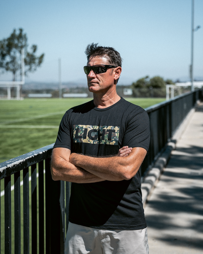 Man wearing a black t-shirt with a logo, standing outdoors with arms crossed.