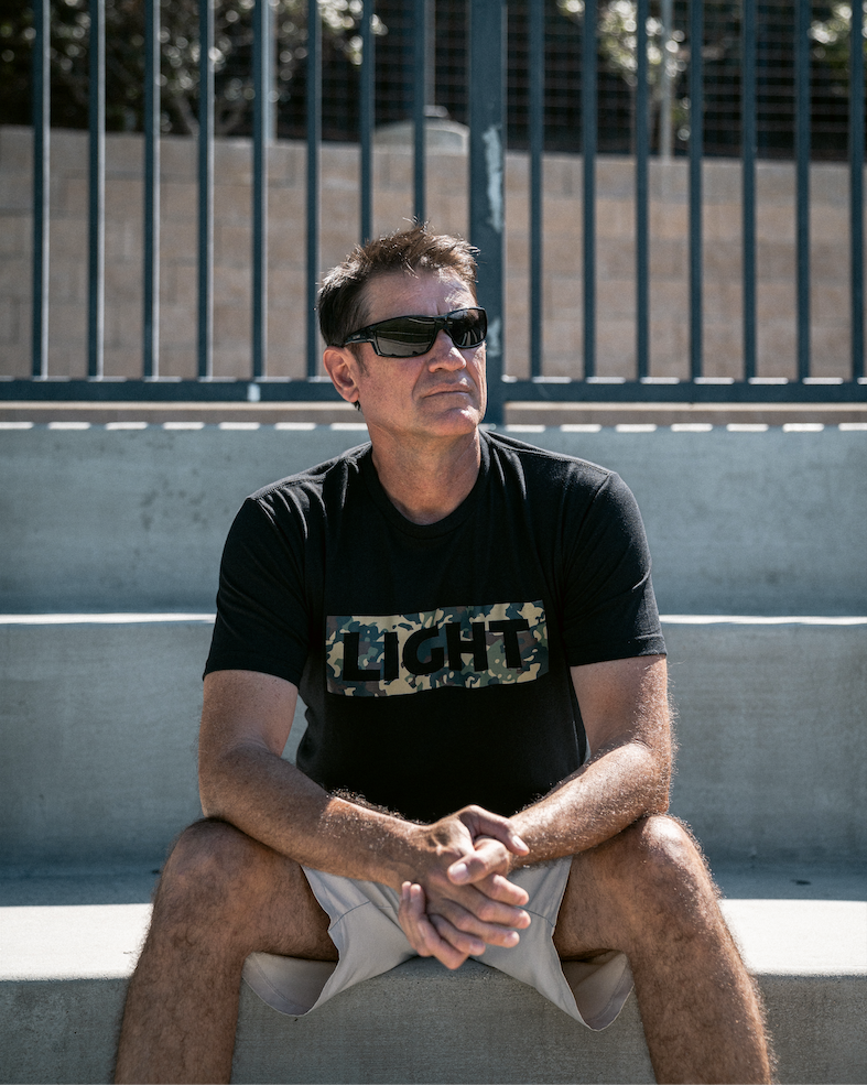 Man wearing sunglasses and a black t-shirt with 'LIGHT' print, sitting on a concrete surface.