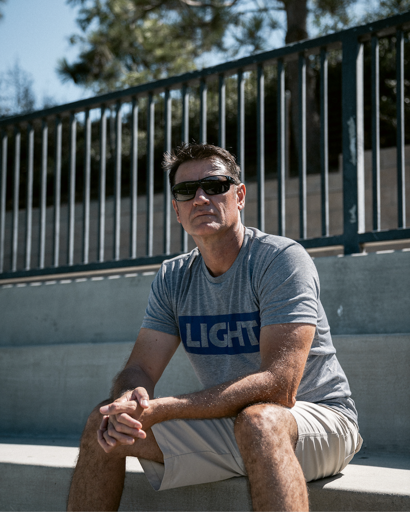 Man sitting on a concrete ledge wearing sunglasses and a gray t-shirt with 'LIGHT' printed on it.