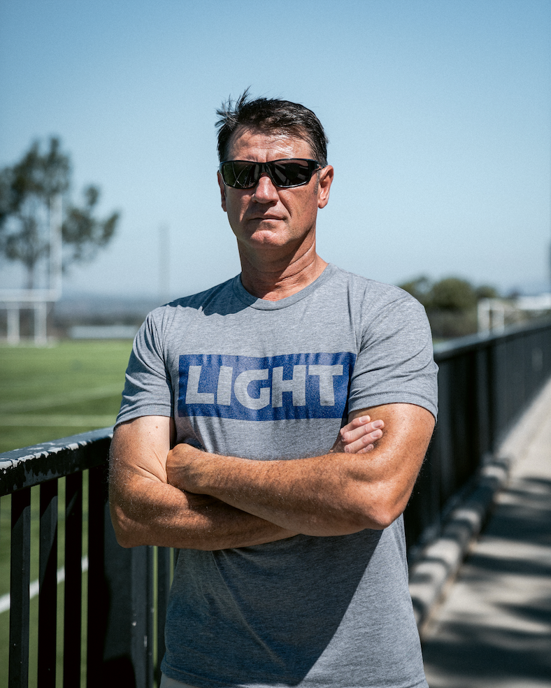 Man wearing sunglasses and a gray t-shirt with 'LIGHT' printed on it, standing outdoors.