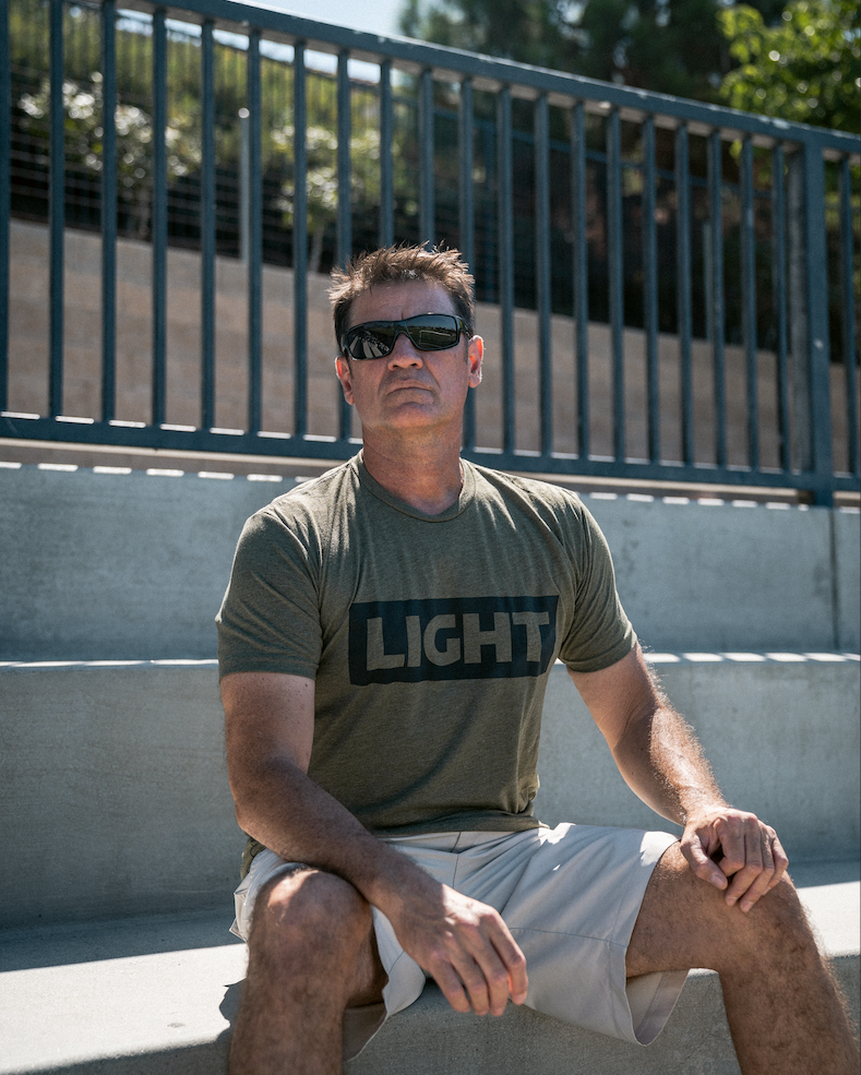Man wearing sunglasses and a green t-shirt with 'LIGHT' printed on it, sitting outdoors.
