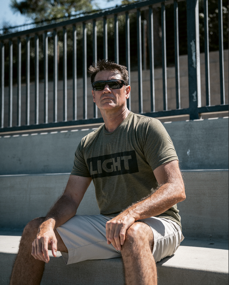 Man wearing sunglasses and a green t-shirt with 'LIGHT' printed on it, sitting outdoors.