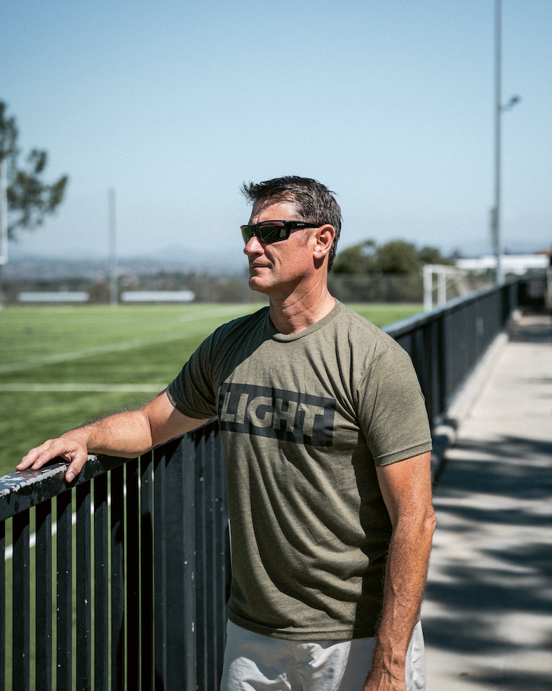 Man wearing sunglasses and a green t-shirt with 'LIGHT' printed on it, standing by a railing.
