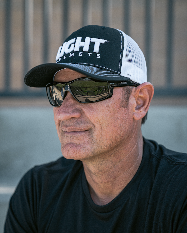 Man wearing a black and white cap with text, sunglasses, and a black shirt against a blurred background.