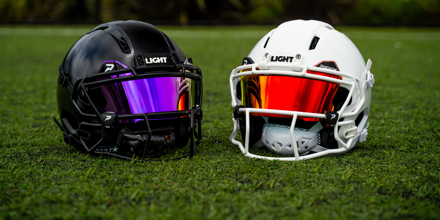 Two LIGHT Gladiator ATK football helmets sitting on a football field with Phenom visors