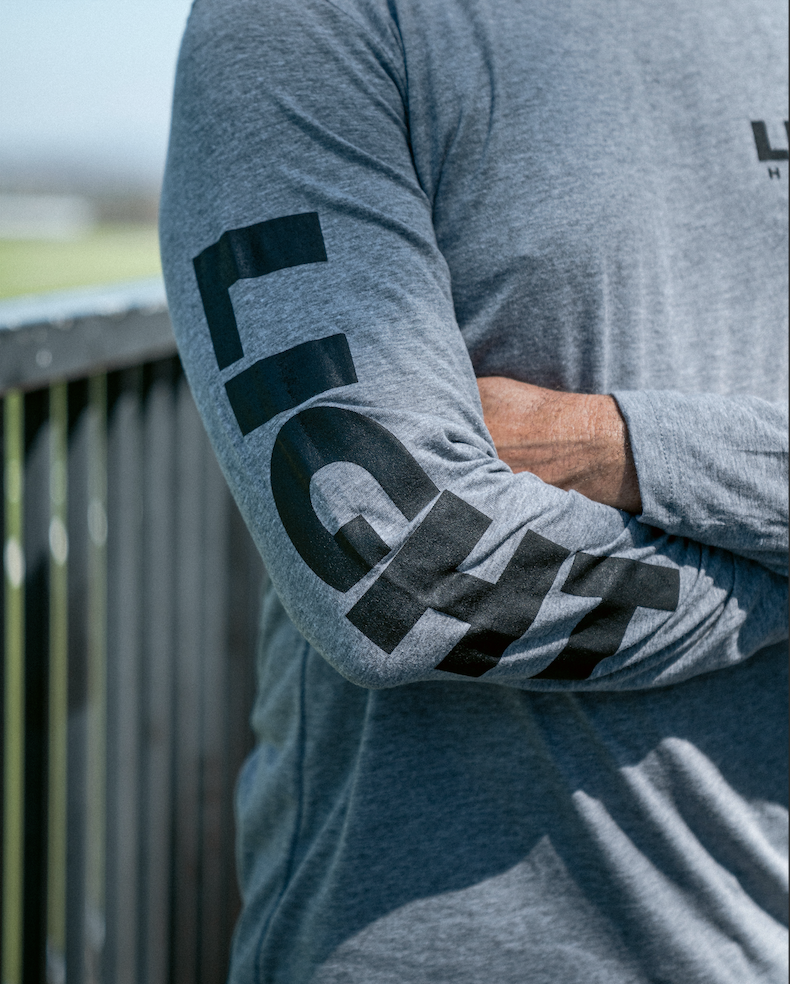 Person wearing a gray long-sleeve shirt with branding on sleeves, standing outdoors.