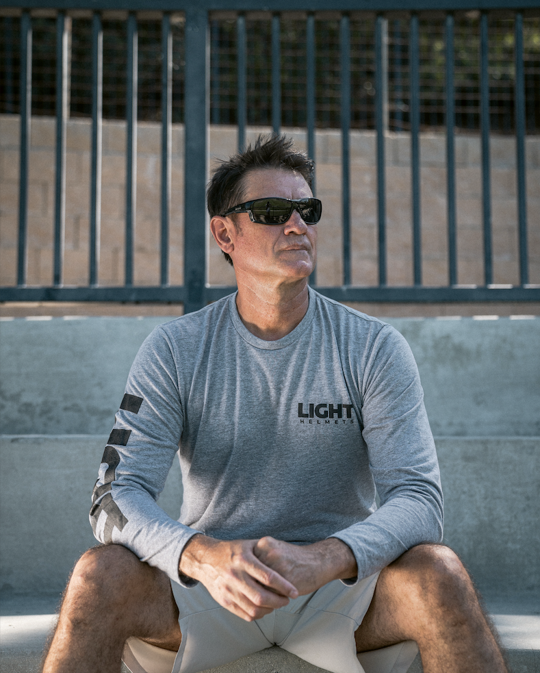 Man wearing sunglasses and a gray long-sleeve shirt with 'LIGHT' logo sitting on a bench.